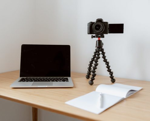 An image of a computer, a DSLR camera on a tripod, and an open notebook