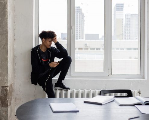 A young black man sitting in the windowsill of a white room, looking at his phone aimlessly